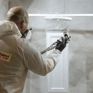 CertaPro Painter in Tyvek suit and mask spraying a hanging kitchen cabinet