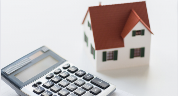 Calculator atop a paper pad next to a pencil and to the left of a small home with a red roof.