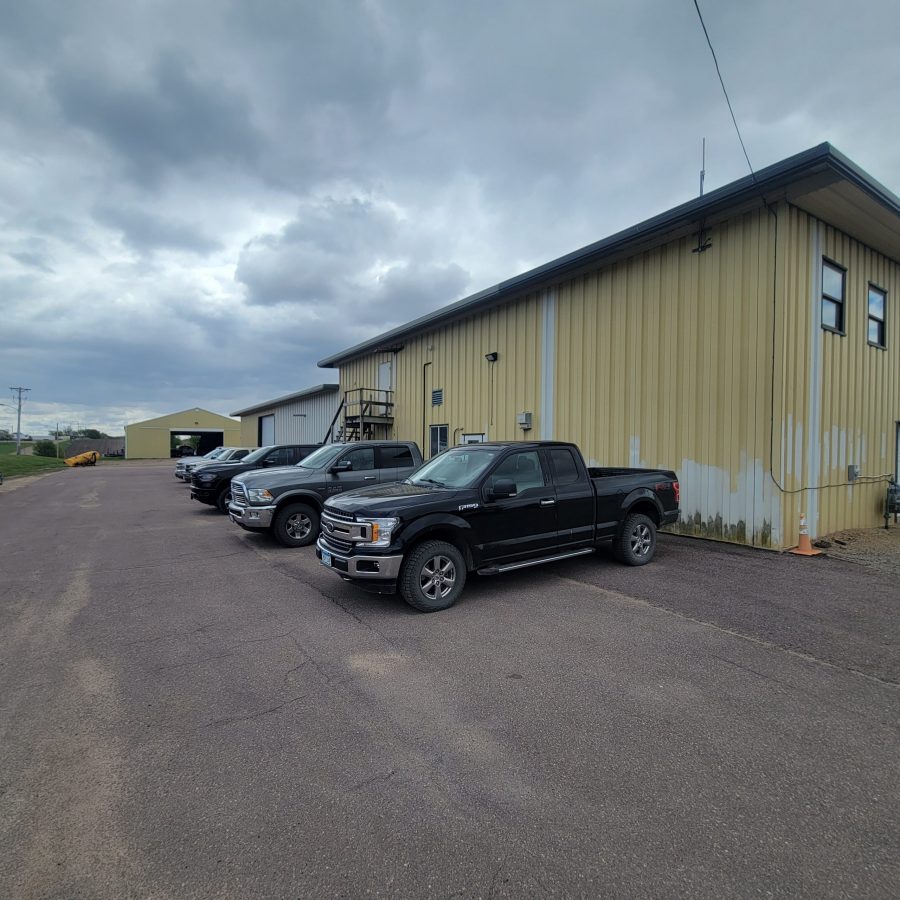 the back view of a greyish yellow paint job done on a commercial building with white doors underneath gloomy weather in Le Center, Minnesota. A few cars are parked behind the building. Preview Image 6