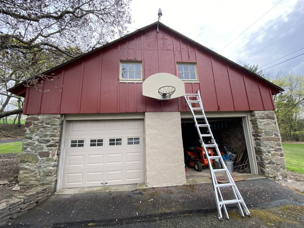 front of garage with basketball hoop, white garage doors, stone and concrete bottom half with top half fresh painted maroon wood