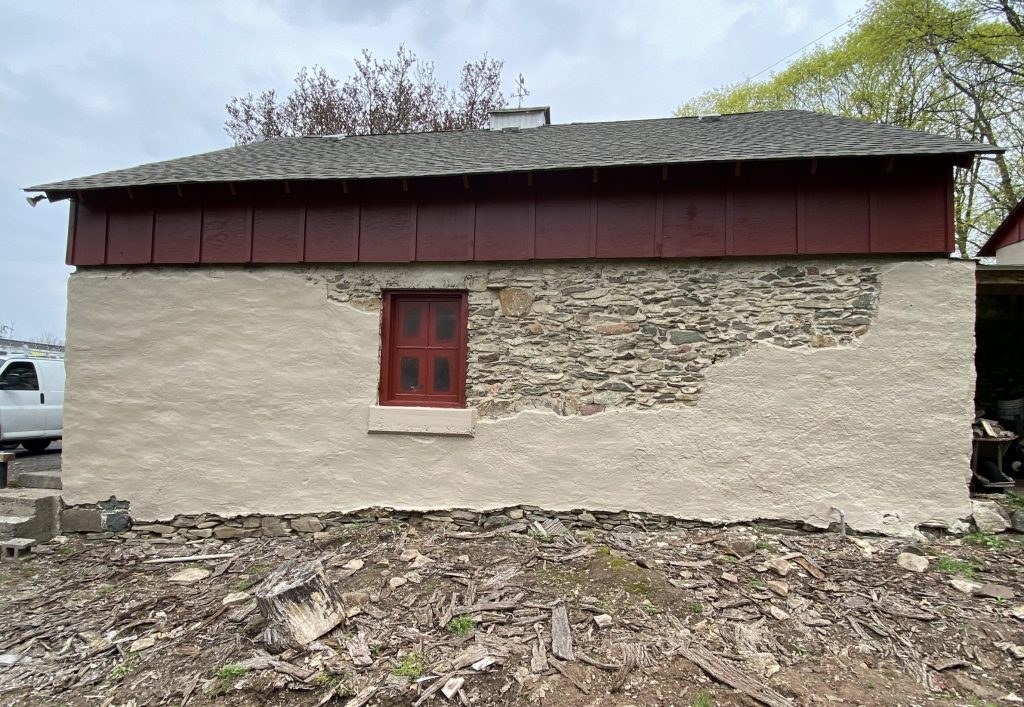 side of garage made of cement and stone painted tan, with maroon trim window and maroon painted wood and grey roof